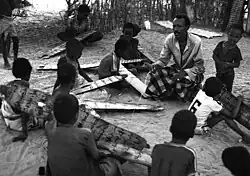 Black and white photograph of a dozen young boys each with a long piece of wood with Arabic writing. They are sitting and standing clustered around a slim middle-aged moustachioed man who is writing bin the sand with a stick and speaking to them. In the background are some bushes with very few leaves