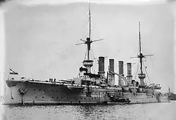 A large warship sits motionless in calm water, with a small boat tied alongside