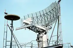 A large gray grid mounted on the top of a ship overlooking a harbor. On the seaport cars are visible, behind the radar the aft end of the frigate is visible as well as a ship docked at the pier.