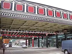 A photograph of a bridge over a road with cars and people on the road. The bridge is built with repeating patterns of red, black, and white.
