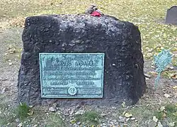 A rectangular, rough-hewn block of stone on the ground. There is a walkway in the foreground, grass and trees in the background. A weathered plaque on the stone reads: "Samuel Adams, Signer of the Declaration of Independence, Governor of this Commonwealth, A Leader of Men and an ardent Patriot."