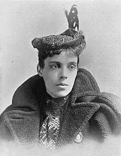 A young white woman wearing a flat cap with a plume, and a bulky coat with wide lapels.