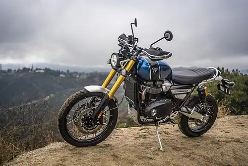 Motorcycle with a blue and black tank, long gold front suspension, and black seat. The motorcycle is parked on its side stand on a dirt road in the mountains, there are houses in the distance.