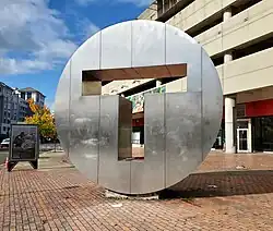 A large metal disk with "T" cut out from the middle, in front of a parking garage