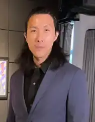 Sean Go, a man with long black hair wearing a dark suit, standing indoors in front of modern artwork.