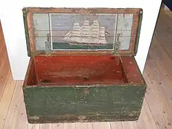 An old looking chest with weathered green paint sits open. There is a decorative painting of a large sailing ship on the inside of the lid with "Auguste Victoria" written under it.