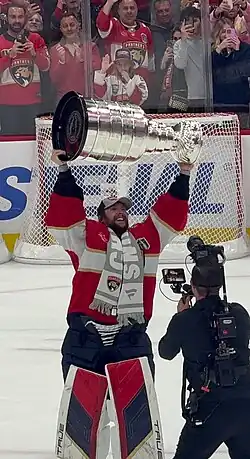 An ice hockey goaltender happily raising the Stanley Cup trophy on the ice. He has a red jersey and a grey scarf.