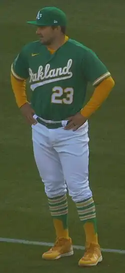 Shea Ryan Langeliers standing on a field with his hands on his hips, wearing the Okland Athletics jersey in green and yellow, a green hat, white trousers and yellow shoes
