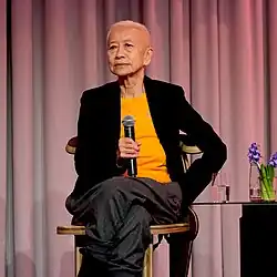 Shu Lea Cheang sitting in a chair on a stage, holding a microphone, looking into the audience