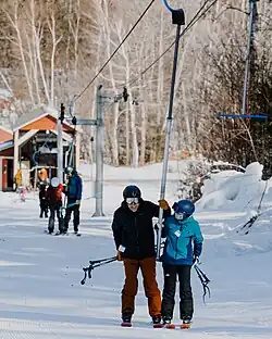 A man and a child ride a T-bar ski lift together at Mt. Abram Ski Area. Both wear helmets and hold onto the lift pole, with snowy trees and skiers behind them on the slope.