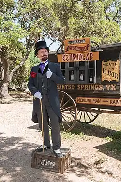 An old-timey man with a long moustache is wearing a suit and tophat and standing on top of a soapbox. Behind him, there is a caravan with adverts describing his snake oil.