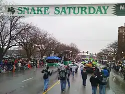 A parade down a street on a cold and rainy day is seen from the middle of the line of parade floats with a banner toward the top of the image saying, "Snake Saturday"