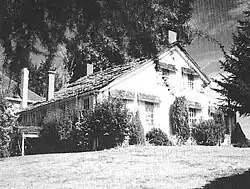 A modest two-story farmhouse with a peaked roof and four chimneys sits under a large tree at the top of a sloping lawn. Bushes and shrubs grow near the house on its two visible sides. A small part of a neighboring house, perhaps only 15 feet (4.6 m) away, can be seen in the background.