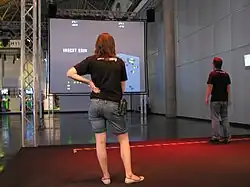 Photograph of two people standing on a open area with a dark carpet. In front of the carpet is a large screen with a Space Invaders-inspired game featuring a large tower being attacked by aliens.