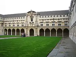 Facade of an ancient building, facing an expanse of grass. At ground floor level the building presents a long colonnade of arches, above which are rows of windows up to a crenellated roof line.