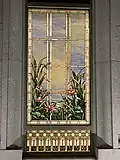 A stained glass window from the Orem Utah Temple, depicting bullrushes and cherry blossoms, next to water elements, with a blue sky with clouds. It is framed by a golden rectangular frame, and has stone masonry surrounding the window.