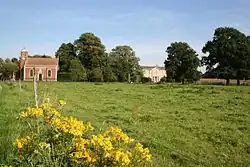A grassy field, its uneaven surface barely discernable, is bright green under a blue summer's sky. In the foreground are the bright yellow flowers of Rattle. A small brick chapel stands in the rear left corner of the field, and beyond that are a row of mature oaks. A gap in the row of trees gives a view of the frontage of Stainfield Hall