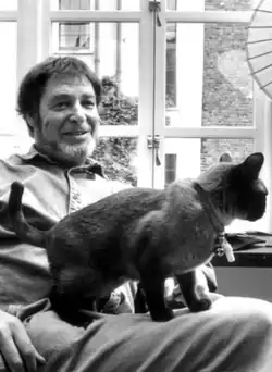smiling bearded caucasian male sitting by a window with a dark British Shorthair crouching on his lap