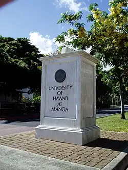 A large stone or landmark made of a marble-like material reads "UNIVERSITY OF HAWAII AT MANOA", with the seal of the university at the top.