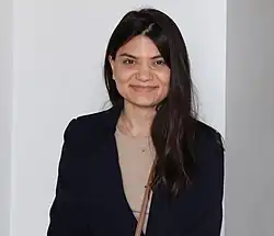 Colour photograph of Susanna Gaboyan aged 26. she is standing wearing a black jacket with a beige shirt, she is smiling widely without showing teeth.