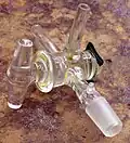 A T-bore glass stopcock in a three way assembly. Two of the outlets end in plain hose adapters while the third ends in a male 14/20 ground glass joint. This stopcock is attached with an easily removed metal spring.