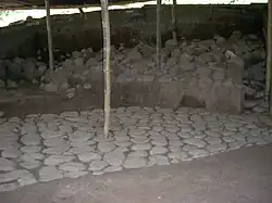 An area of excavation under a corrugated roof supported by wooden poles. The foreground shows an even cobbled pavement running diagonally from left front to right back, where it disappears into a vertical wall of partially excavated rubble and soil.