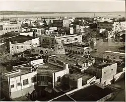 Sepia-toned helicopter view of a city with roads flooded by water and buildings damaged. Another helicopter can be seen near the middle of the image.