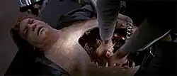 A white male lies on a table seemingly unconscious. His torso is opened from chest to stomach in the formation of a mouth with sharp teeth along the edges. A doctor attempting to revive him has both his hands inside the exposed, empty cavity.