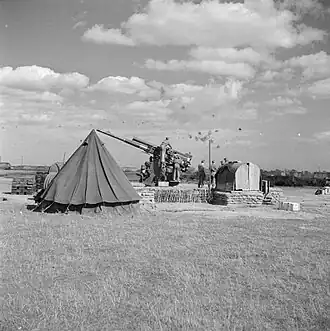 Black and white photo of a 3.7-inch anti-aircraft gun with ack-ack bursts in the sky above.