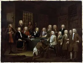 A painting of six men wearing white wigs, and dressed elegantly, seated around a table covered with a green cloth. Eight men, also wearing wigs and dressed in finery, are standing behind the table. One man without a wig, dressed in a rougher style, and with a rough-looking face, stands at the front of the table, addressing the others. Another rough-looking man stands at the back. A black man wearing only a cloth around his waist kneels in front of one of the men wearing a wig. The black man is wearing an iron device around his neck. Another iron device is lying on the floor behind him.