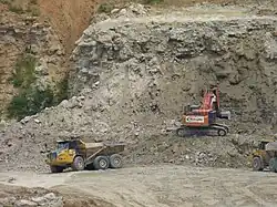 A photograph showing quarrying operations with a mechanical digger in the centre extracting rock from a cliff face. To the left a lorry is just leaving with a full load of rock and to the right another lorry is waiting to collect its load from the digger.