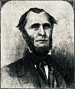 A man with dark, receding hair and a dark beard wearing a white shirt and dark jacket