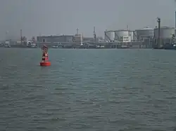 A red port-side buoy with an oil wharf and tankers in the background