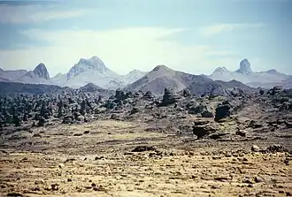 A hazy-looking yellow field of desert sand with rocks in the foreground and dark mountains and the blue sky in the background