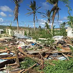 A daytime photograph of a completely destroyed building with debris near palm trees and other damaged buildings