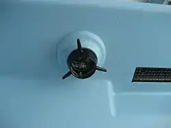 A moulded bulge proturding from the underside of a mounled plastic surface; at the base of the bulge, a short wide hollow black plastic cylinder with three narrow, outward-tapering radial spokes. Inside the cylinder, two black crossbars and a cotter pin hold the black plastic part on; behind them, a grater with spiral blades scrapes across the square surface of a block of solid soap.