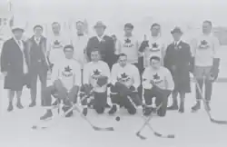 Black and white photo of a hockey team outside on natural ice, including four men each dressed in a dark overcoat, and nine players dressed in hockey equipment white sweaters with a maple leaf crest