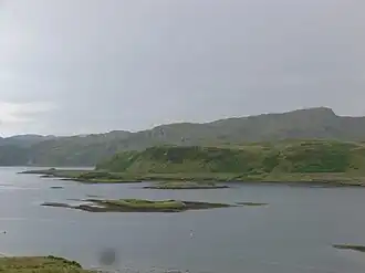 The north of Torsa with visible raised beach line below the cliffs. Sgeir a' Bhodaich is at centre and Glas Eilean between there and Torsa. The heights of Dùn Crutagain on the mainland of Scotland are beyond.