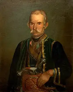 A black-and-white portrait of a man with a stern expression and a well-groomed moustache, dressed in ornate traditional clothing with intricate embroidery and a decorated belt. He holds a gun tucked into his belt, symbolising authority.