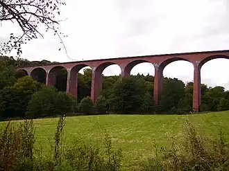 A low angle looking up view of a brick viaduct with woods on the left and a skyline above the viaduct
