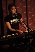 A woman plays keyboard and sings in front of a red patterned background.