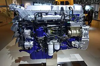 Volvo D13 engine on a display stand at an industry trade show. The blue engine block contrasts with various white accessories, black hoses and wires, and a silver engine valve cover