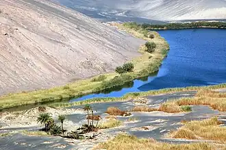 Blue lake surrounded by green-yellow vegetation, within a desert and at the foot of a hill