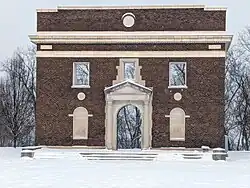 A brick facade to a demolished building stands in the snow, with trees seen through the empty door and windows.
