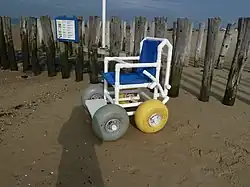 A chair with four wide, balloon-style wheels is in front of a fence at the beach
