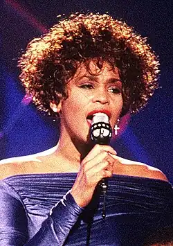 Whitney Houston performing on stage, wearing an off-the-shoulder, deep blue gown with ruched fabric. Her short, curly hair frames her face, and she holds a microphone while singing.