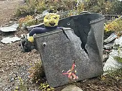 A memorial site on a hillside features a sign that reads, “Riding to Remember Wichita State University Football Team, October 2, 1970.” A yellow jersey with the number “12” hangs from a tree trunk, and a white cross is planted nearby. Wreckage from the plane is scattered across the area, with a bouquet of sunflowers and a jacket placed as tributes. Trees and fallen logs are visible throughout the scene.