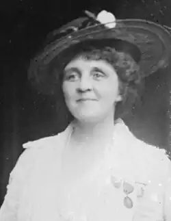 A smiling white woman with curly hair. She is wearing a hat with a wide brim, a light colored jacket, and medals pinned to her jacket.