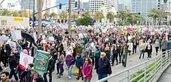 Marchers with signs walk down a street from right to left. Buildings and palm trees stand in the background.
