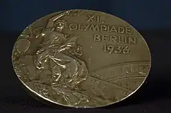 A tarnished gold medal featuring a person in a toga, and the words "XI Olympiade Berlin 1936".